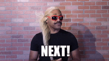 Celebrity gif. Wearing a long blonde wig, a moustache, and red sunglasses, Robert E Blackmon stands in front of a brick wall. With both hands, he points from right to left and gives us a playful: Text, "Next!"