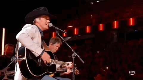 ACM Awards gif. Male musician in cowboy hat is seated as they strum a guitar while sing into a standing mic with a smile on their face as the camera rotates around the singer, they pull away from the mic to grin