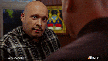 TV gif. Wearing a gray plaid shirt, Joe Minoso as Joe from Chicago Fire gives a surprised reaction to someone in the foreground, then tilts his head slyly.
