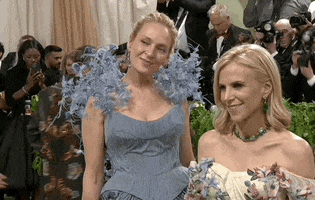 Met Gala 2024 gif. Uma Thurman poses with Tory Burch, wearing one of her designs, a powder blue gown with full pleats, structured bodice, flowers growing tall out of her shoulders.