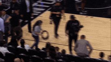 Sports gif. Manu Ginóbili, a former player on the San Antonio Spurs shows up to watch a game and he waves at the audience with a grin on his face.