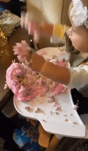 Video gif. Baby sitting in a high chair with a pink birthday cake smashed on their tray. They wave their arms with excitement and turn towards us with a happy expression, holding out their hands filled with cake and cake all over their face. 