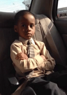 Video gif. A young boy sitting in the back of a car with his arms crossed looks at us and rolls his eyes in annoyance.