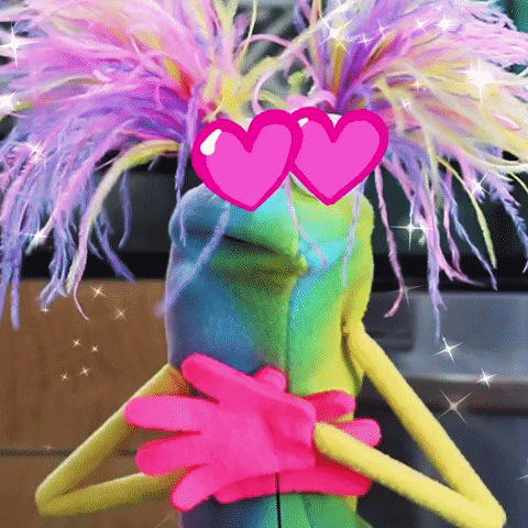 Video gif. Green puppet with hearts for eyes and fluffy neon eyelashes presses its pink hands to its chest, overcome with love.