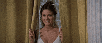 Movie gif. Mary-Margaret Humes as Miriam in History of the World: Part 1 is hiding between two curtains looks excited as she tiptoes with glee and holds both hands with crossed fingers up.