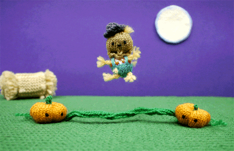 Stop motion gif. Two crocheted pumpkins swing a jump rope in a field beneath a full moon as a scarecrow, hosts, and three-headed monster take turns jumping in a continuous loop. When the three-headed monster jumps, it loses one of its heads, then stops to pick it up.