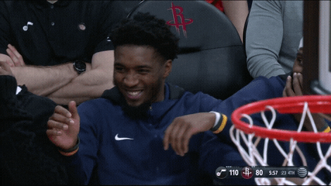 Sports gif. Donovan Mitchell from the Utah Jazz is cracking up on the bench with his teammates. He laughs so hard that he nearly falls out of his seat, and he lays against his teammates who are all laughing just as hard.