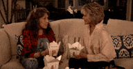 TV gif. Lily Tomlin as Frankie and Jane Fonda as Grace from Grace and Frankie sit facing each other on a white couch. They toast with two open cartons of Chinese food as if saying "cheers!"