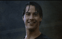 Movie gif. Keanu Reeves as Johnny Utah in Point Break smiles and gives a cool thumbs-up while standing soaking wet from the pouring rain around him.