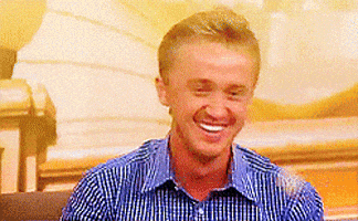 Celebrity gif. Tom Felton is being interviewed but something makes him laugh big time and he covers his mouth with both hands as he cracks up.