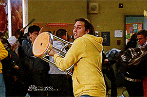 TV gif. The cast of Community is in a school and chaos is breaking out. A man grabs a stool and runs over to a window, tossing the stool through the glass.