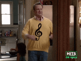 Sponsored GIF. Eric Stonestreet is in a kitchen and shimmies the reveal of his new sweater donning a treble clef symbol. Camera zooms in as he continues to shimmy and declares with pride, "Here comes treble!"