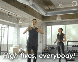 Video gif. Walking towards us in a gym, a woman puts her hands in the air, giving us high fives. She says, “High fives, everybody!”