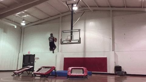 Sports gif. Six basketball players take turns jumping off of a trampoline and passing each other the ball by bouncing it off of the backboard. The seventh player jumps the highest and weaves the ball between his legs before finishing with a slam dunk that shatters the glass.
