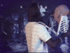 Video gif. Man wearing a werewolf mask is slow dancing with a skeleton that has a Golden Retriever mask on.