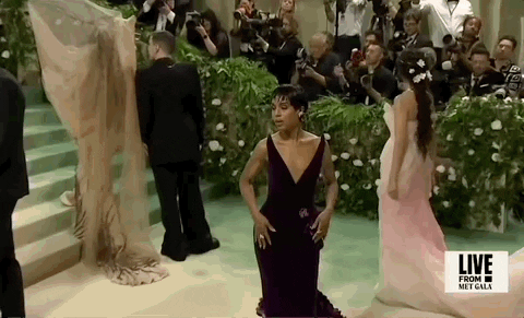 Met Gala 2024 gif. Kerry Washington, in a maroon velvet gown with a structured plunging neckline that frames her face, poses for the cameras, mugging moodily, landing with her hand elegantly primping the nape of her neck.