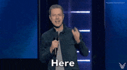 Video gif. Presenter Geoff Keighley on stage at the 2024 Summer Game Fest. He is wearing a dark blue button-down shirt over a light gray t-shir. He speaks into the microphone he holds in his right hand as his left hand gestures to emphasize the first word of his announcement, "Here," followed by, "is the first look.", " The announcement is displayed as a caption at the bottom of the gif.