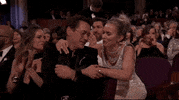Oscars 2024 GIF. Emily Blunt leans over the row to put her arms around Robert Downey Junior and share a laugh with him.