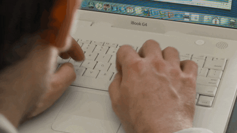 Video gif. Over the shoulder view of a man typing on a vintage Mac laptop and we see his hands from his perspective as they clack away.