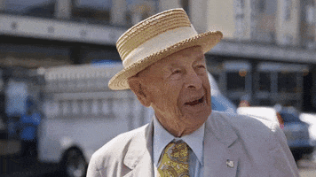 Video gif. Old man wearing a straw boater hat turns his head from the side, facing us, squinting with a smile.
