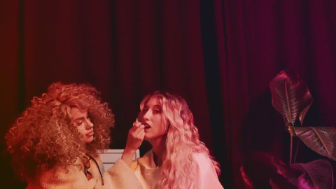 Music video gif. From Louis the Child's video for "Evalyn," a blonde wavy-haired woman sings while a brunette curly-haired woman touches up her make up, sitting in front of a red curtain next to a plant.