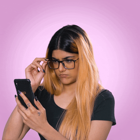 Celebrity gif. Singer Ananya Birla adjusts her glasses while looking at something surprising on her cell phone.