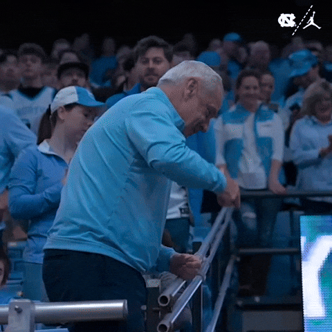 Sports gif. Roy Williams, coach of the North Carolina Tar Heels, points and smiles as he leans sideways over a railing. He turns back, laughing and applauding while fans surround him in the stands.