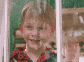 Movie gif. Macaulay Culkin as Kevin from Home Alone waves out a window as snow falls outside.
