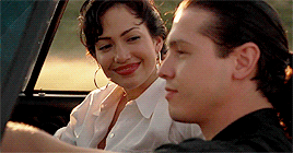 Movie gif. From the passenger seat, Jennifer Lopez as Selena smiles at Jon Seda as Chris Perez and wraps her arms around his shoulders and kisses him while he drives in a top-down convertible.