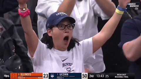 Sports gif. A woman wearing a UConn hat looks shocked with wide eyes and mouth gaping as she holds her arms up. Then she rests them on her head and looks around while people clap in the stands behind her.