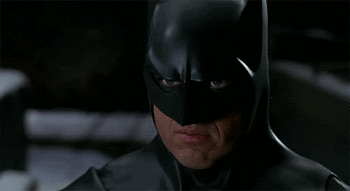 Movie gif. Michael Keaton as Batman. He's wearing his full Batman suit and smiles slowly.