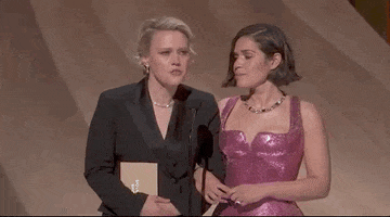 Oscars 2024 GIF. Kate McKinnon and America Ferrera stand on stage and McKinnon leans into the microphone to inquire with utter seriousness, "To whom have I been sending my tasteful nudes?" Ferrera turns to us with a blank deadpan expression.