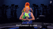 Video gif. Jess King rides a Peloton dressed in a costume of Mrs. Frizzle from The Magic School Bus. She does arm stretches while pedaling and says, “Halloween is one of my favorite holidays and I’ve been having so much fun.”