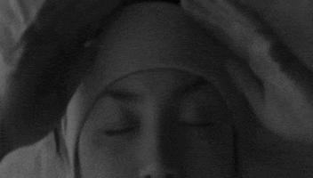 Video gif. Woman is lying in bed wearing a headband pulls it down over her eyes, revealing the message "let me sleep."