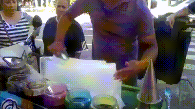 Video gif. A man with a food cart carves a big block of ice and shovels it into a cone shaped mold. He takes the mold off and has created a cone shaped snow cone on a stick. He then pulls spoons out of jars of thick syrup, letting the syrup drip back into the jars. He twirls the snow cone under the syrup to create a swirled design on the cone. He repeats this a few times with different colored syrups and hands the snow cone off once it’s completely covered. 