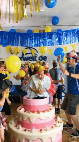 Video gif. Group of people in wild party outfits energetically dance in a room decorated with blue and yellow party decorations. A man in a gold hat has arguably the most enthusiastic dancing as he stands inside a multi-tiered cake statue with his mouth wide open like he's feeling the moment and dancing just for him. 