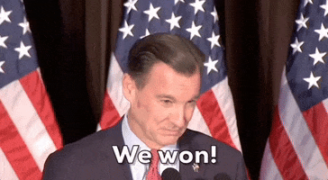 Political gif. Thomas Suozzi stands at a podium in front of American flags and says, “We won!" while cameras flash around him. 