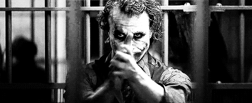 Movie gif. Black and white clip of Heath Ledger as The Joker in The Dark Knight. He stands in a jail cell clapping rhythmically as he glares down at us.