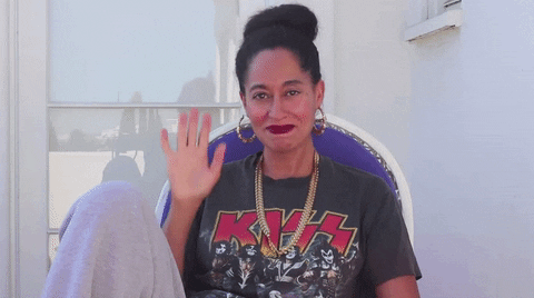 Celebrity gif. Tracee Ellis Ross sits in a chair with her knee up while wearing a kiss band t-shirt. She looks at us with a smile and waves.
