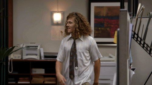 TV gif. Blake Anderson as Blake in Workaholics misses a high five as he struts through an office with a carefree smile. Text, "TGIF" 