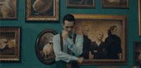 Music video gif. Brendon Urie is standing in a hallway filled with picture frames and he looks down, upset at  something. He sighs deeply and crosses his arms in contemplation.