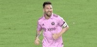 Sports gif. Soccer player Leo Messi wearing a pink Adidas jersey smiles with an open mouth, holding out his hands and running across the field toward someone.