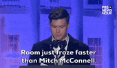 Video gif. Saturday Night Live's Colin Jost stands at a podium at the 2024 White House Correspondents' Dinner and delivers a joke, saying "Room just froze faster than Mitch McConnell." He momentarily breaks into laughter, then stops as he puts his hand against his temple in disbelief at his own joke.