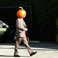 Video gif. A man wearing a suit and carrying a briefcase walking through a parking lot with a Jack-O-Lantern on his head.