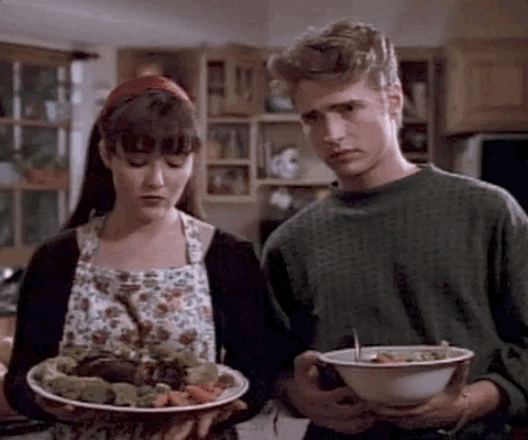 TV gif. Jason Priestly as Brandon Walsh and Shannon Doherty as Brenda Walsh from Beverly Hills, 90210 appear slightly guilty as they fidget uncomfortably and avoid contact while holding platters of Thanksgiving food.