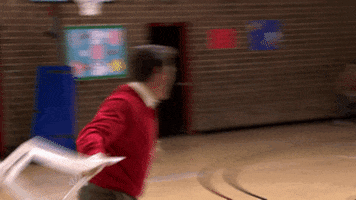 Parks and Recreation. In a basketball gym, Nick Offerman as Ron throws a white plastic lawn chair across the court toward the bleachers where people are sitting.