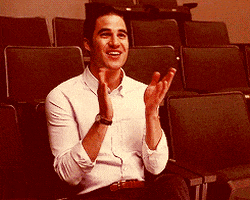 TV gif. Darren Criss as Blaine in Glee claps as he rises from his seat in an auditorium.