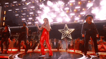 TV gif. Country singer Lainey Wilson performing live on stage at the 2024 ACM Awards in bright red pants. She sings passionately into a microphone.