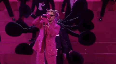 Oscars 2024 GIF. Ryan Gosling is in the middle of his performance for I'm Just Ken and he puts the microphone back on the stand. The microphone stand is way too tall and Gosling shortens it, bringing it down to his height. 