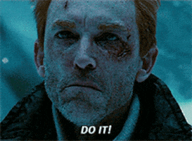 Movie gif. A man with a wound on his cheek quivers with anger and then yells, “Do it!”